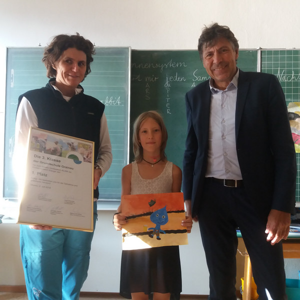 Eine Frau mit Siegerurkunde, ein Mädchen mit selbst gemaltem Bild und ein Mann stehen in einem Klassenzimmer